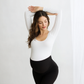 Pregnant woman wearing black maternity leggings and white long-sleeve top stretching with arms raised.