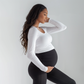 Pregnant woman wearing black maternity leggings with a supportive high-rise waistband and a white long-sleeve top against a plain light background.